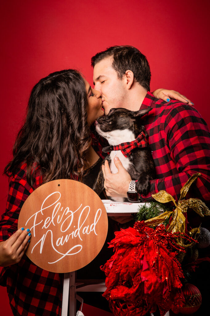 Foto en fondo rojo estudio de Navidad, pareja con french bulldog y letrero de Feliz Navidad escrito en caligrafía