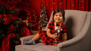 Niña pequeña sentada en poltrona gris sosteniendo un cascanueces con fondo rojo navideño